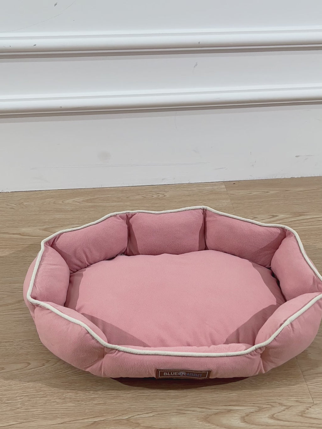 Pink pet bed on a wooden floor with a visible brand label.